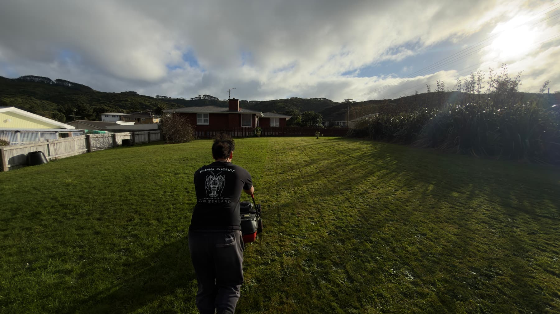 A Taiao Services crew member mowing a Porirua section under a bright, cloudy sky
